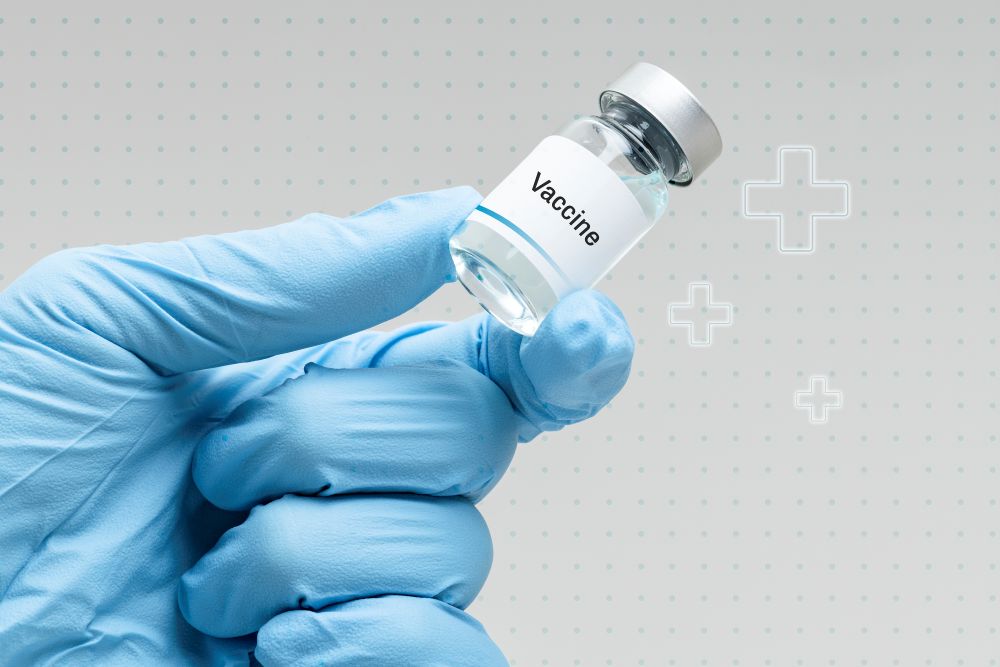 HPV: Anvisa autoriza uso de vacina na prevenção de cânceres além do colo do útero 6 Close de uma mão com luva cirúrgica azul segurando um pequeno frasco de vidro de vacina contra um fundo cinza claro com ícones de cruz médica.