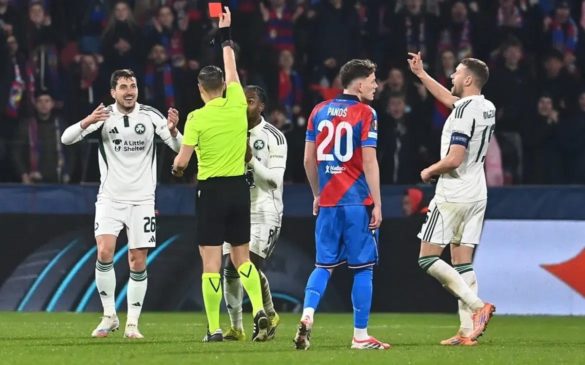 Árbitro mostra cartão vermelho para jogador enquanto atletas protestam em campo de futebol lotado