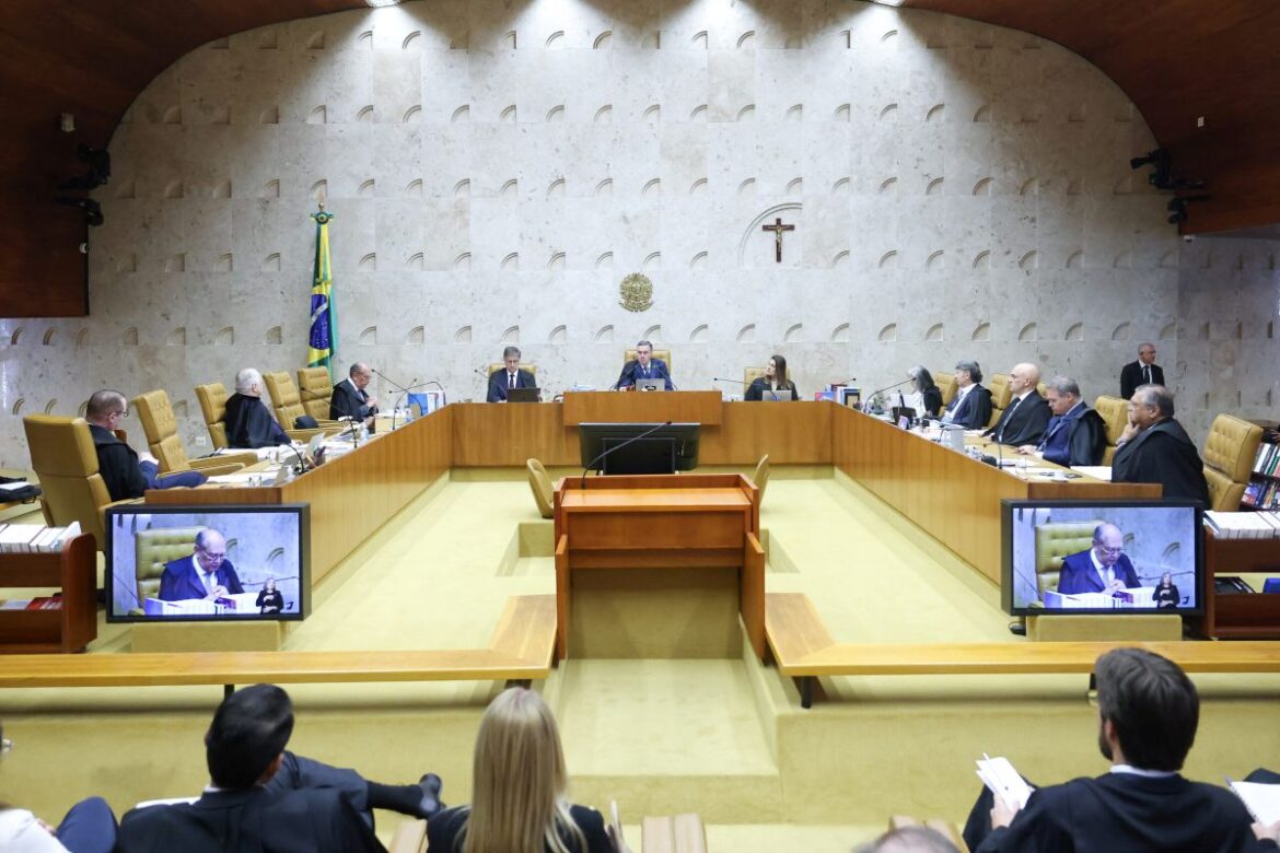 Ministros do Supremo Tribunal Federal (STF) em sessão plenária, sentados em uma mesa de madeira em formato de U, com a bandeira do Brasil e o crucifixo ao fundo, e telas exibindo alguns ministros.