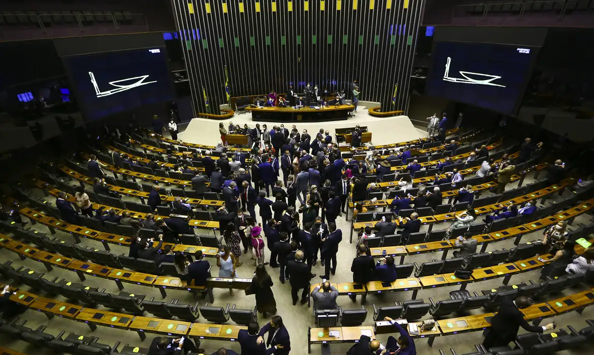 Fim da escala 6x1: Governo deve enviar projeto após o Carnaval 6 Visão panorâmica do Plenário da Câmara dos Deputados em Brasília. Deputados em ternos circulam entre as bancadas de madeira organizadas em semicírculos. Ao fundo, a mesa diretora sob um painel de ripas verticais e dois grandes telões exibindo ícones de votação.