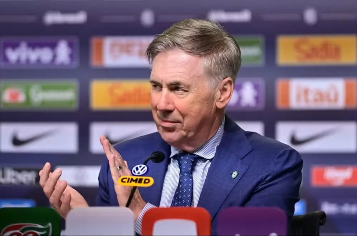 Treinador de terno azul gesticula em coletiva com microfones e painel de patrocinadores ao fundo