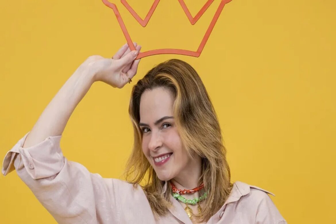 Ana Paula sorrindo segura coroa de papel laranja sobre a cabeça em fundo amarelo