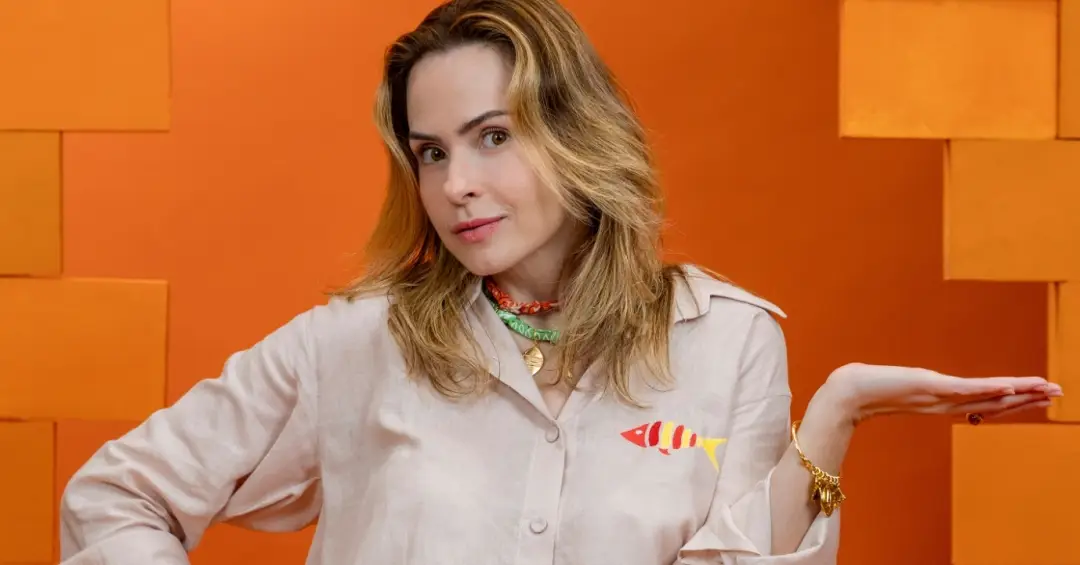 Ana Paula Renault vence a prova do líder e fala sobre plano ousado para o paredão 6 Ana Paula faz gesto com a mão em fundo laranja, usando camisa clara e acessórios coloridos