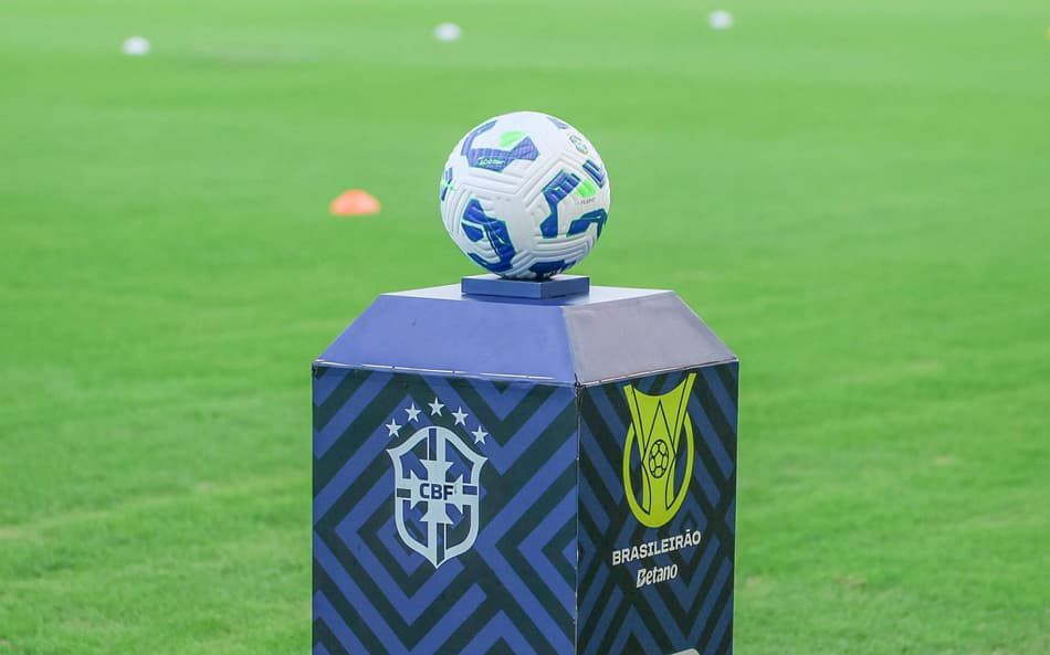 Brasileirão 2026: palpites da rodada, onde assistir e melhores apostas 6 Bola de futebol branca com detalhes azuis posicionada sobre pedestal do Brasileirão 2026 em campo verde