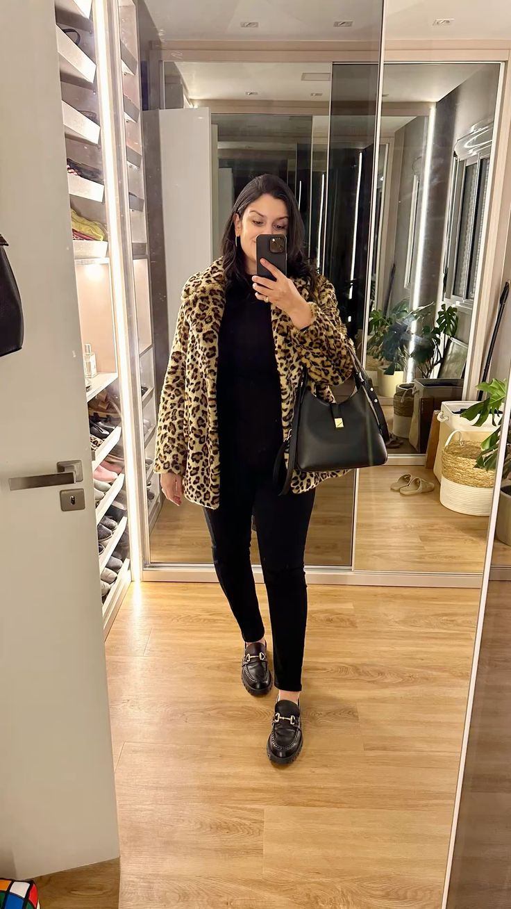 Moda Outono 2026: confira as principais tendências 8 Foto em frente ao espelho de uma mulher vestindo um casaco de pelúcia com estampa de onça (animal print) sobre um look preto total, usando mocassins tratorados de couro e segurando uma bolsa de ombro preta estruturada.