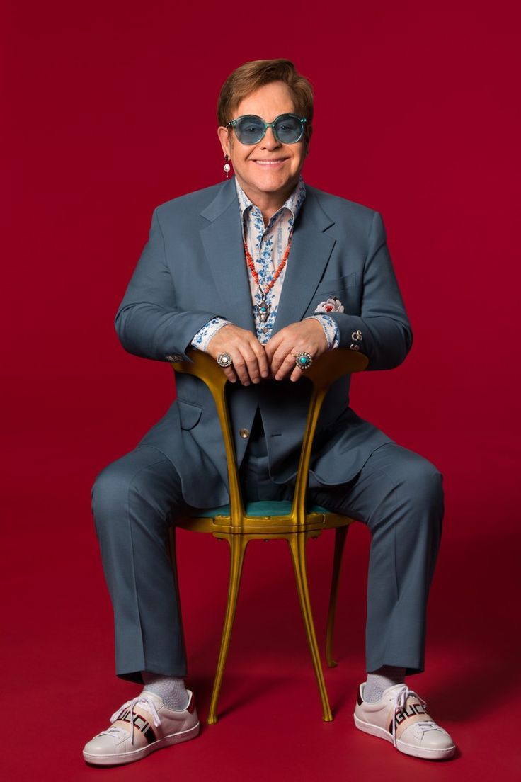 Rock in Rio 2026: veja como ficou o line-up com as novidades recentes 6 Elton John sorrindo, sentado em uma cadeira dourada com as costas para a frente, em um fundo vermelho vibrante. Ele usa um terno azul-petróleo, óculos de sol azuis, joias exageradas e tênis brancos.