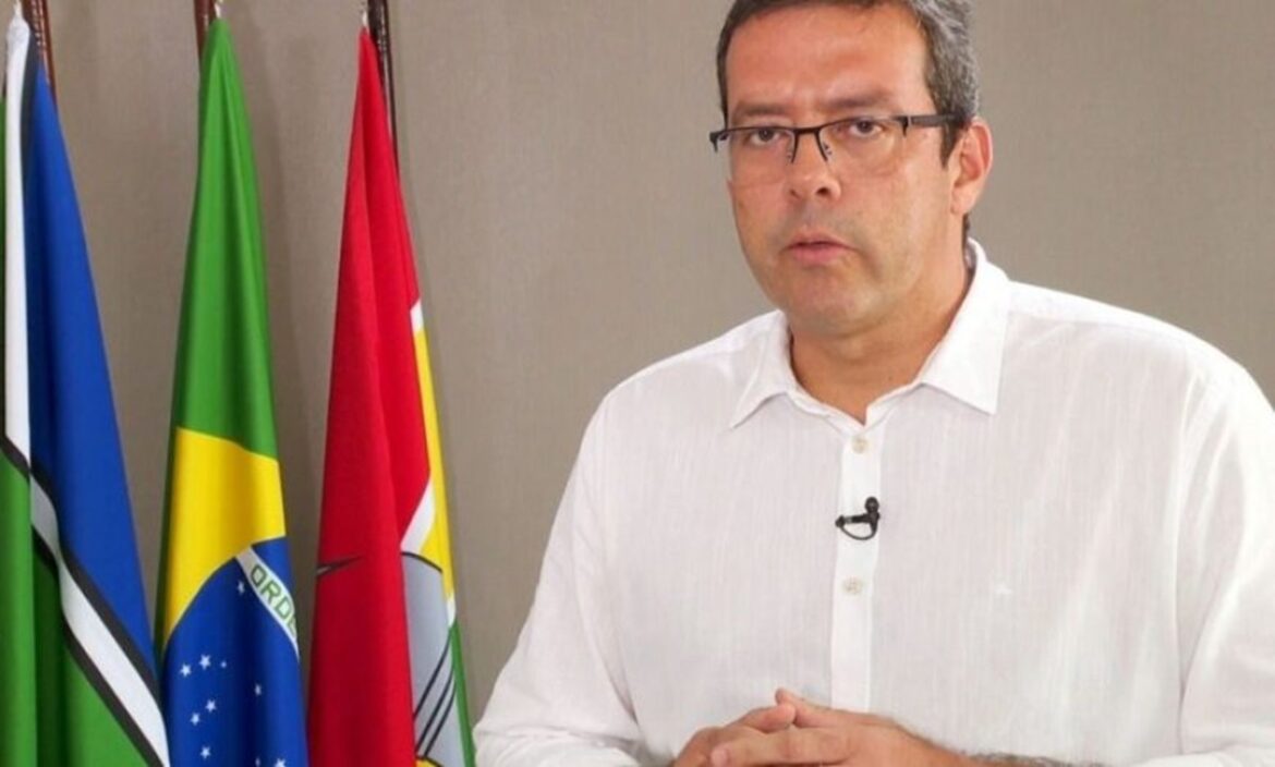 Homem de óculos com camisa branca fala em frente a bandeiras do Brasil e do município