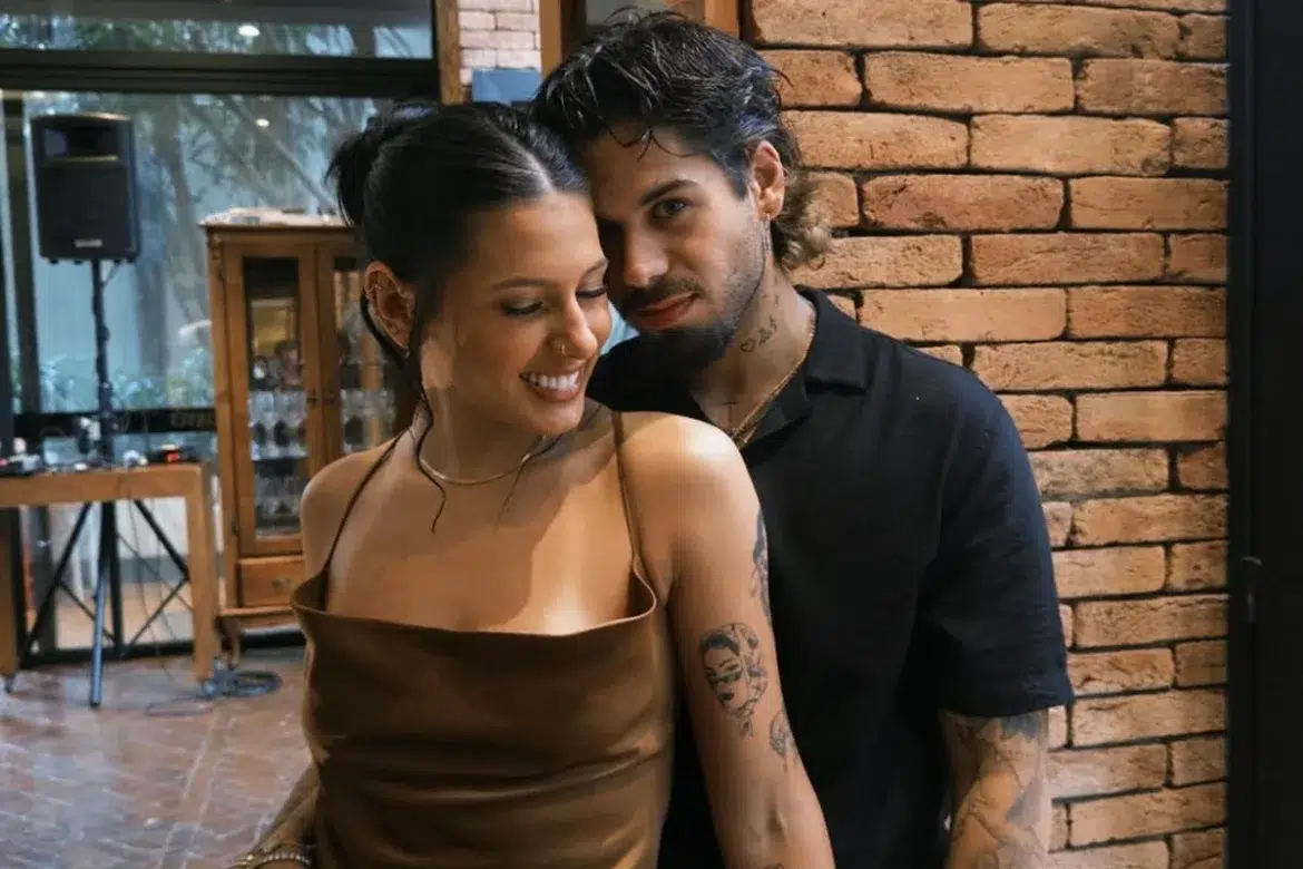 Ana Castela e Zé Felipe sorrindo em um ambiente rústico com parede de tijolos.