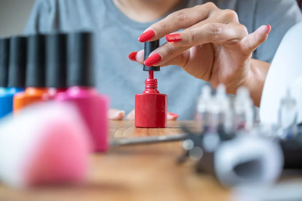 Mulher com unhas vermelhas abrindo frasco de esmalte em gel sobre mesa de madeira