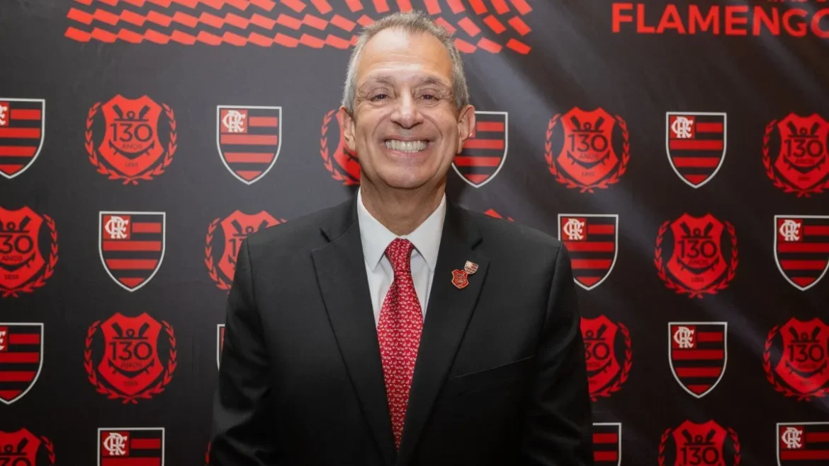 Homem de terno escuro e gravata vermelha sorri diante de painel com escudos do Flamengo
