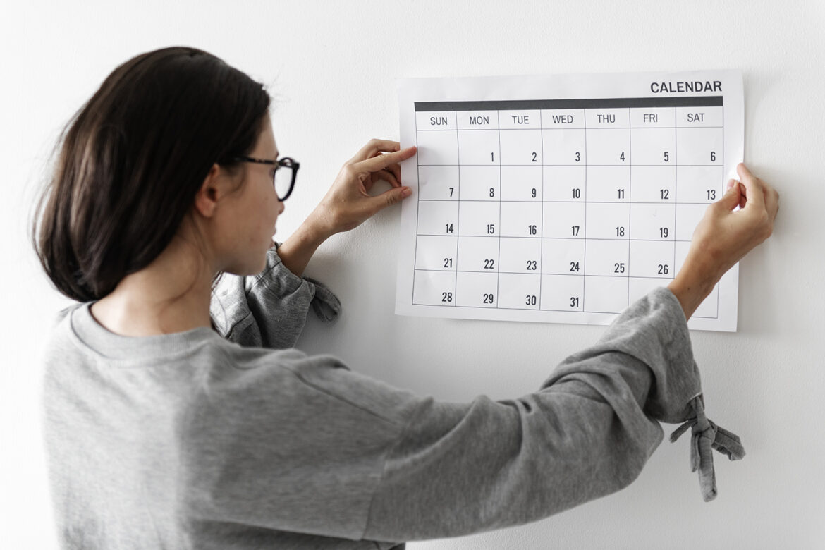 Mulher de óculos ajustando calendário na parede enquanto observa datas do mês