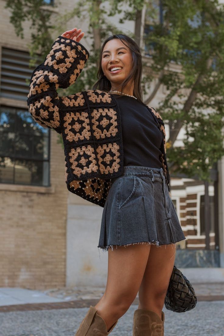 Moda Outono 2026: confira as principais tendências 7 Mulher asiática sorrindo, posando ao ar livre. Ela usa um casaco de crochê com quadrados pretos e beges, blusa de gola alta preta, saia jeans cinza de cintura alta e botas de cano alto beges. Ela segura uma bolsa preta.