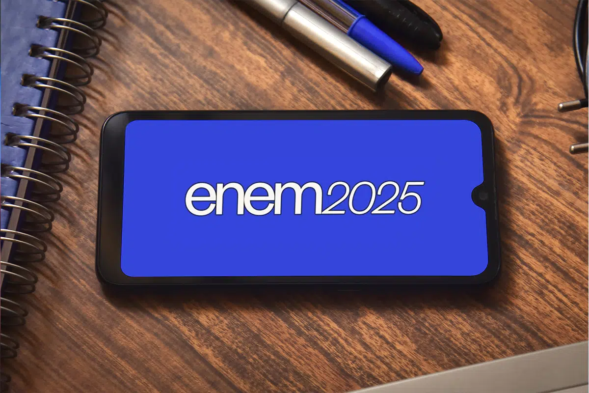 INEP lança plataforma para emissão de certificado de conclusão do ensino médio 6 Celular sobre mesa de madeira exibindo o logo do Enem 2025 em fundo azul, representando a nova plataforma de certificação digital do ensino médio.