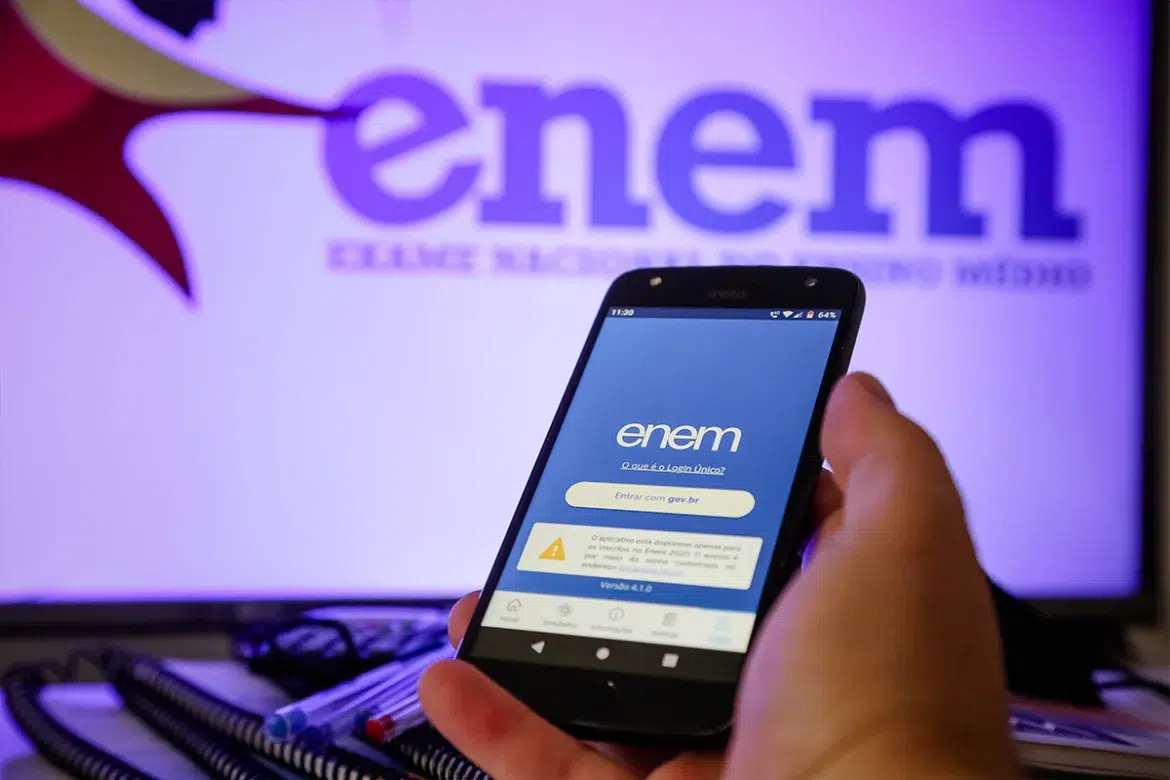 Mão segurando celular com a tela de login do aplicativo do Enem e botão de acesso via Gov.br, com monitor exibindo a marca do Enem ao fundo.