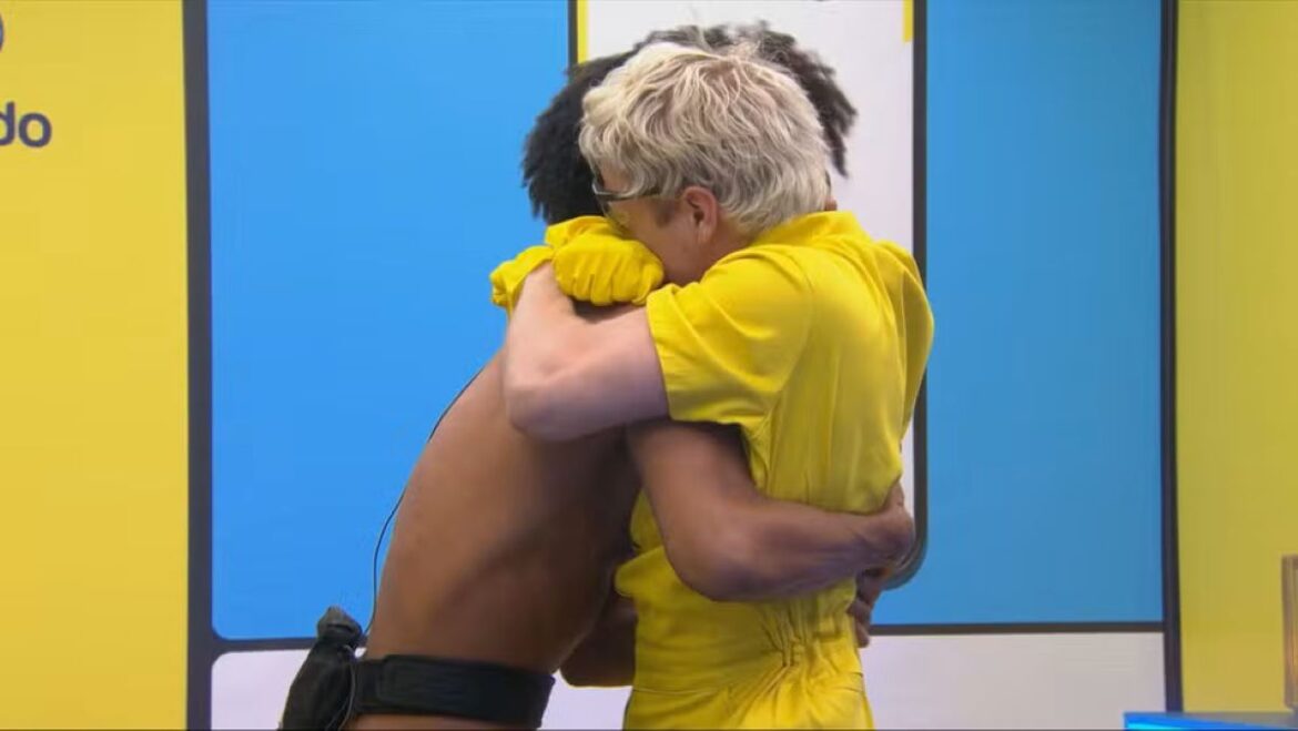 Juliano, vestindo um macacão amarelo, abraça Breno emocionadamente após sua entrada no Quarto Secreto do BBB 26, em frente a um painel azul e amarelo.