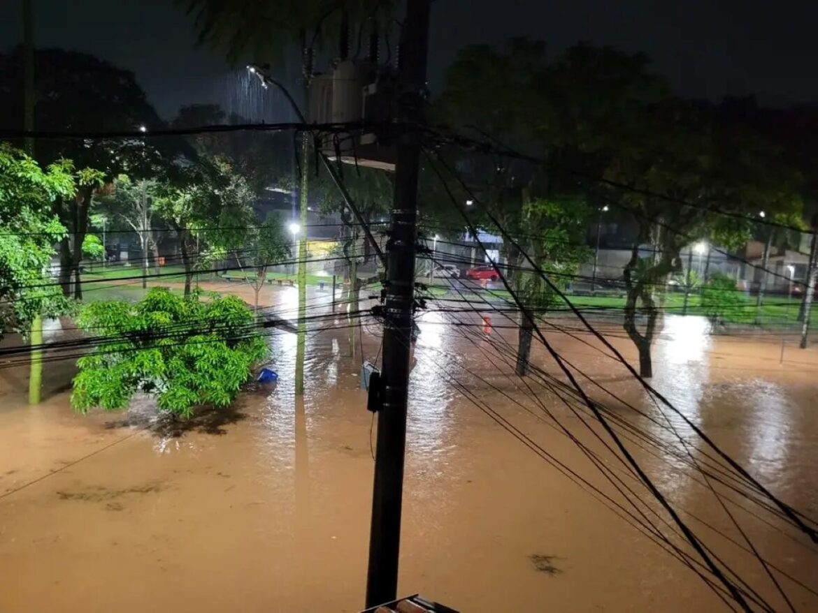 Vista aérea noturna de uma praça urbana inundada em Juiz de Fora; a água barrenta cobre o gramado e as calçadas sob a luz dos postes.