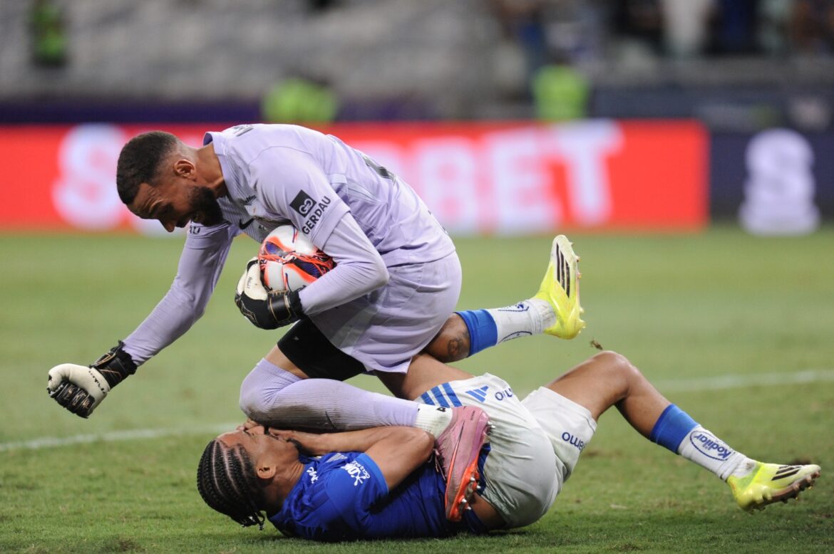 Goleiro segura bola enquanto atleta adversário está caído no gramado durante partida acirrada