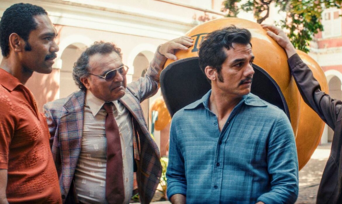 Cena do filme "O Agente Secreto" com Wagner Moura em destaque, vestindo camisa azul e olhando para o lado, acompanhado por outros três atores perto de um orelhão amarelo vintage.