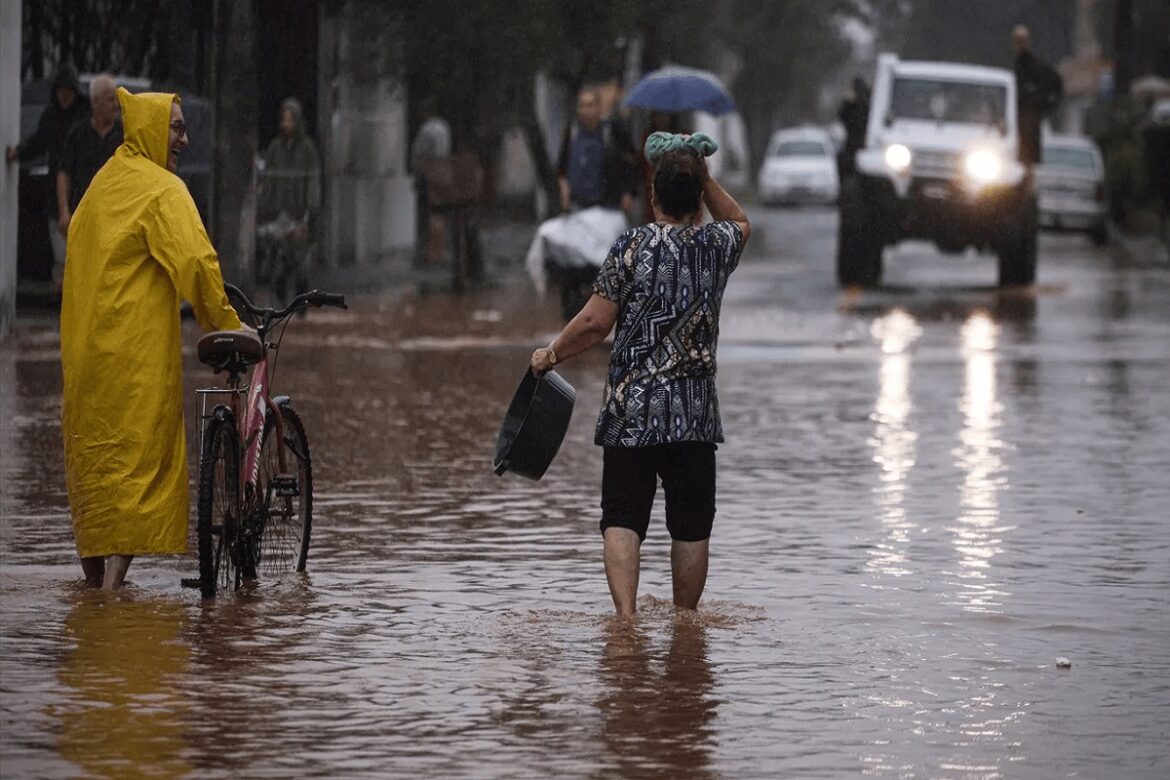 Pessoas caminham por rua alagada, uma empurrando bicicleta de capa amarela, outra carregando balde sob chuva intensa