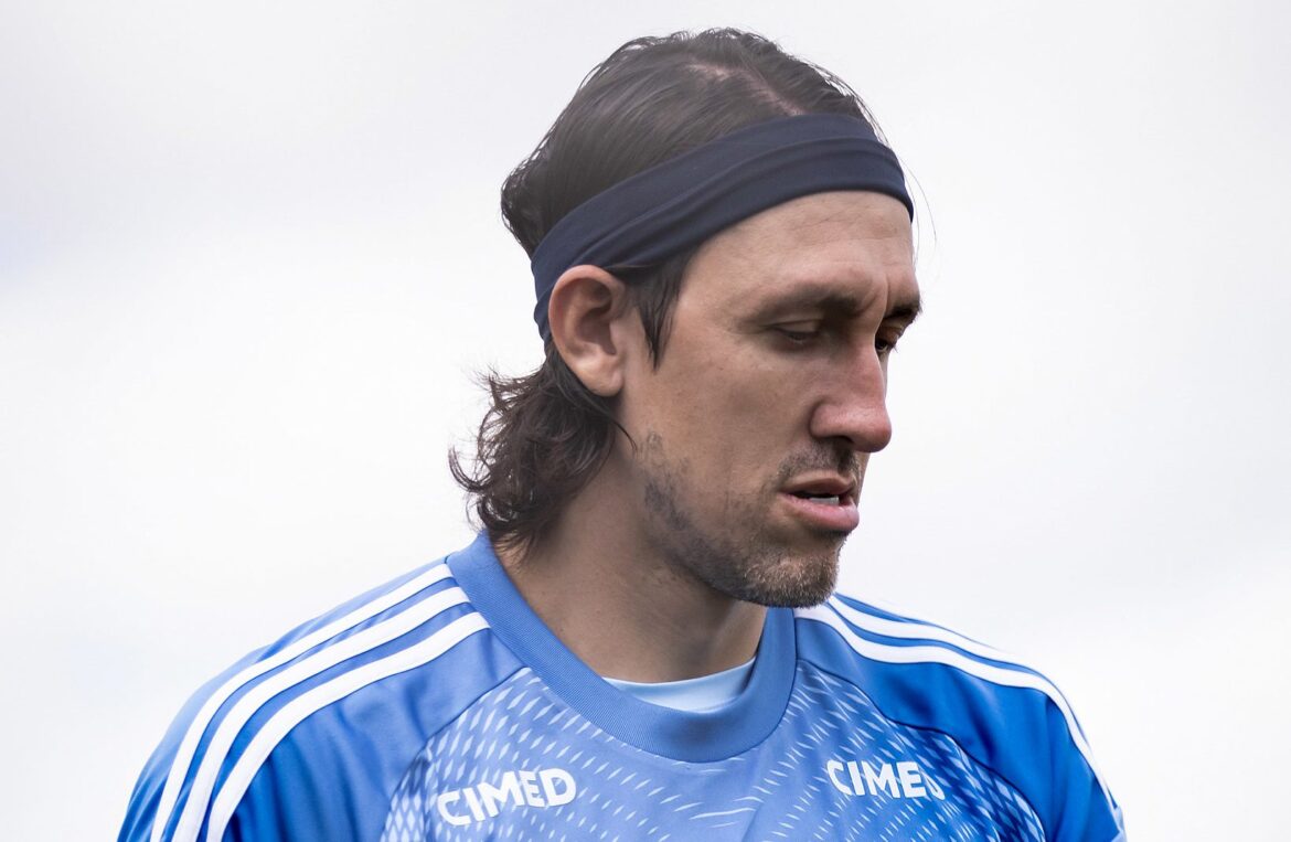 Atleta de uniforme azul olha para baixo com expressão séria em ambiente externo