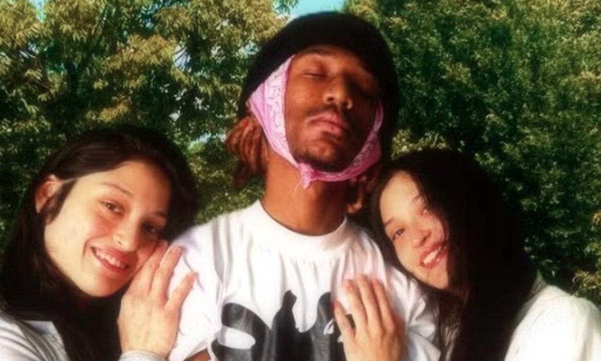 Duas gêmeas sorrindo abraçadas ao mesmo homem em parque arborizado