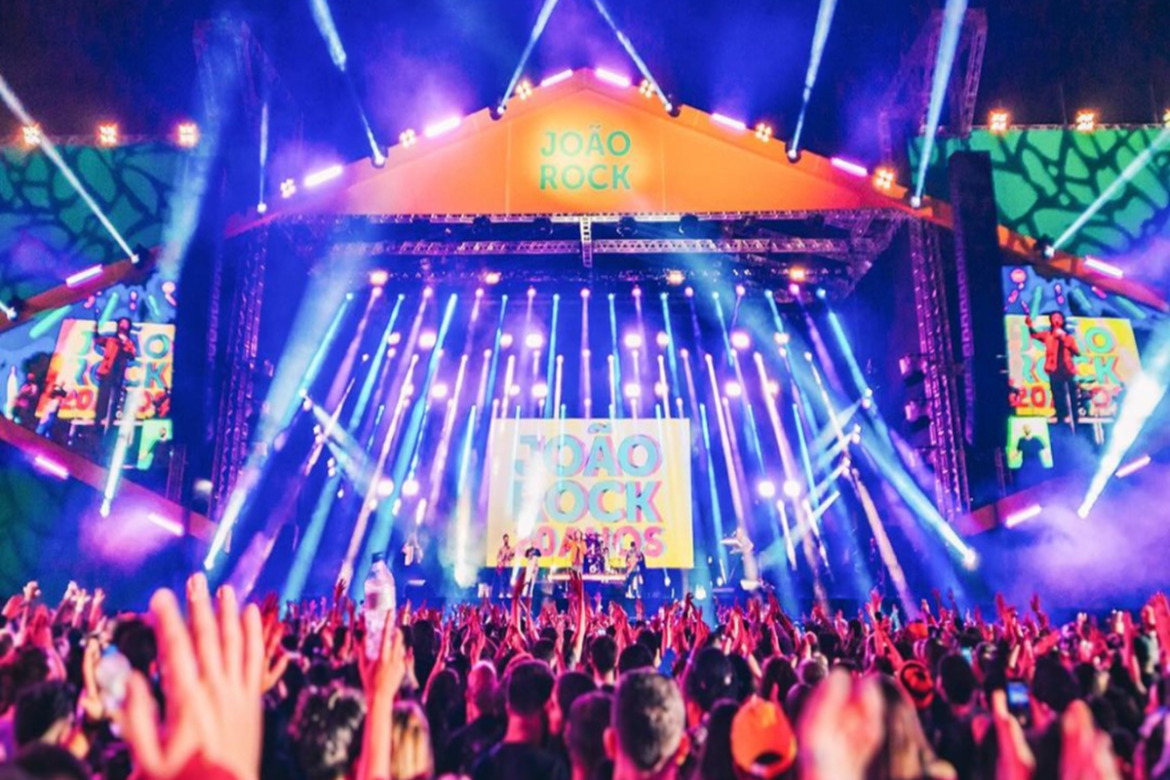 Multidão com mãos levantadas assiste a apresentação de banda sob luzes coloridas e palco iluminado