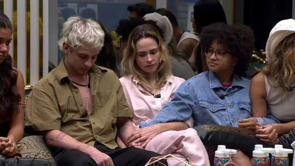 Enquete BBB 26: Quem você quer que saia no 10º paredão? Vote agora 6 Grupo de participantes sentados, mãos dadas, expressão séria em sala durante noite de eliminação