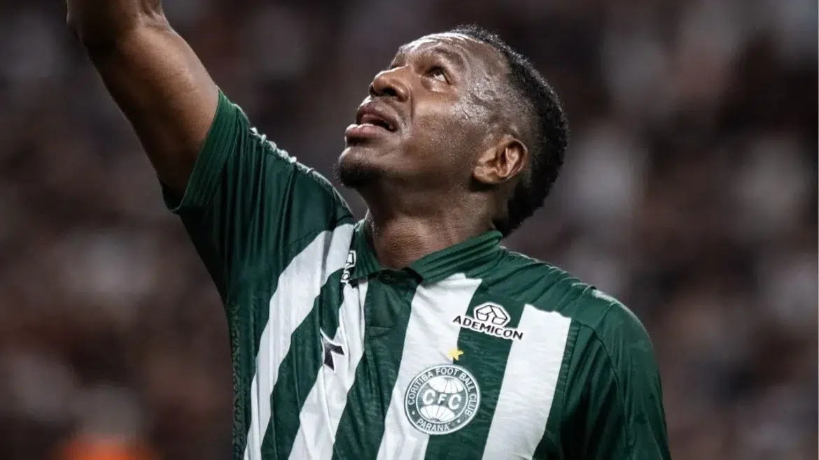 Atleta com uniforme do Coritiba ergue o braço e olha para cima, expressão de frustração