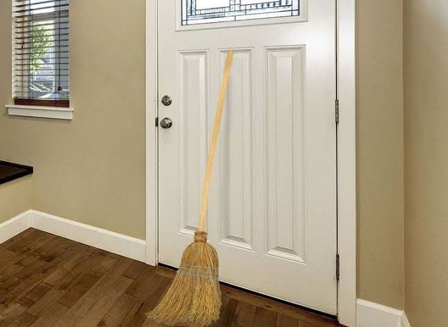 O truque da vassoura atrás da porta funciona? A psicologia explica por que o gesto realmente faz a visita ir embora 6 Vassoura de palha apoiada no centro de uma porta branca fechada em um ambiente interno moderno com piso de madeira.