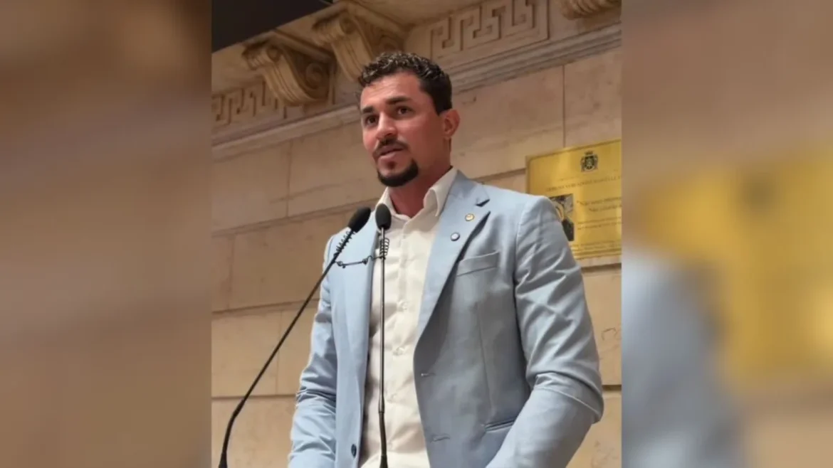 Vereador em pé falando ao microfone em plenário com parede de mármore ao fundo