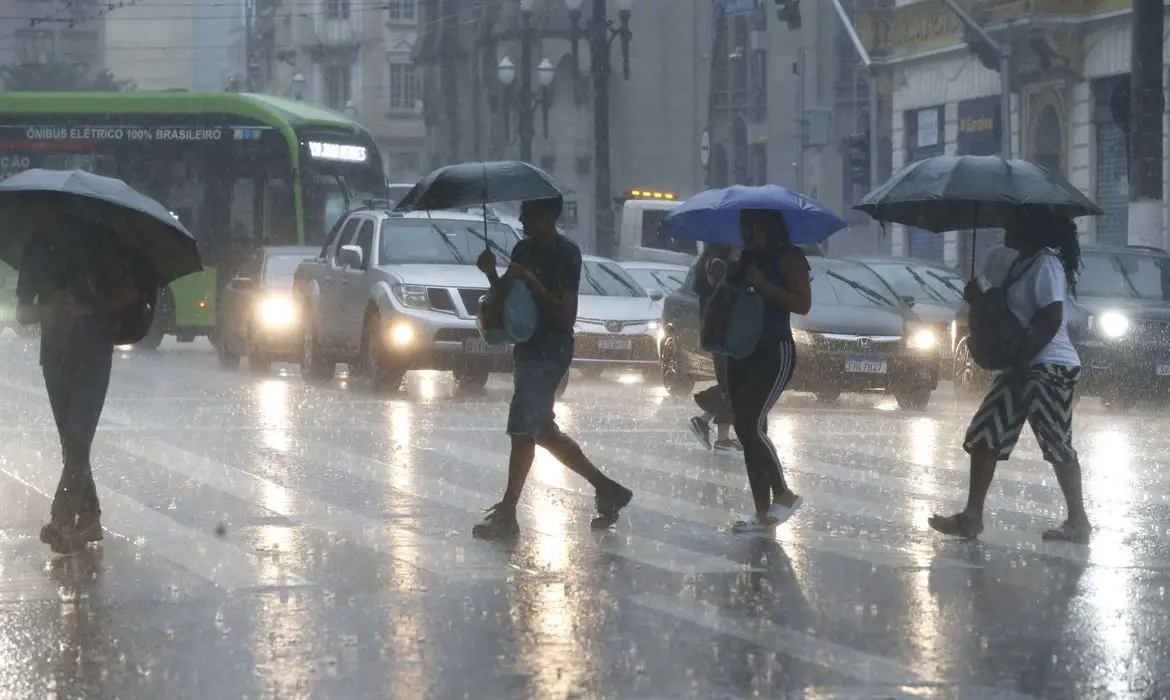 Alerta para 5 estados: frente fria traz risco de chuva forte hoje (28) e amanhã (29) 6 Pedestres com guarda-chuvas atravessam rua molhada sob chuva intensa, com veículos e ônibus elétrico verde ao fundo