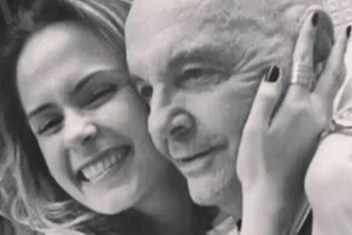 Mulher loira sorrindo abraça homem idoso careca, Gerardo Renault, de 96 anos , em foto preto e branco