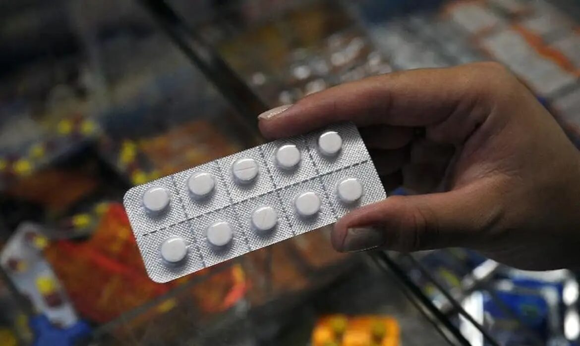 Mão enluvada segura ampola de dipirona monoidratada 500mg/mL com caixas do medicamento Hypofarma sobre a mesa.
