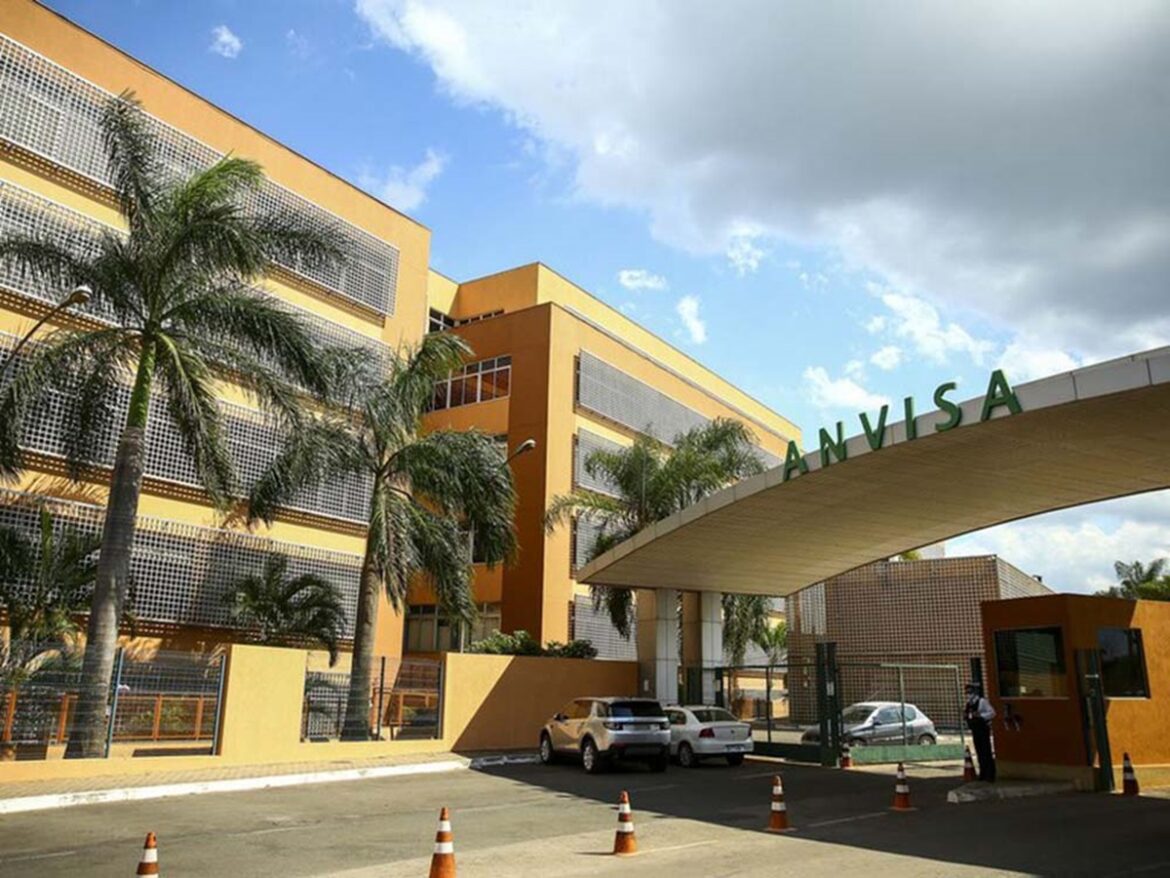 Fachada da sede da Anvisa em Brasília com letreiro verde, edifício amarelo, palmeiras e portaria de acesso