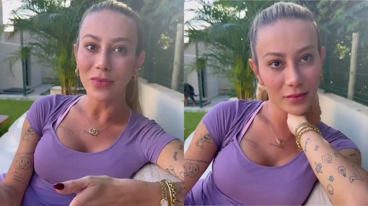 Apoline em blusa lilás com tatuagens nos braços, usando colar e pulseira dourados, em área externa com jardim