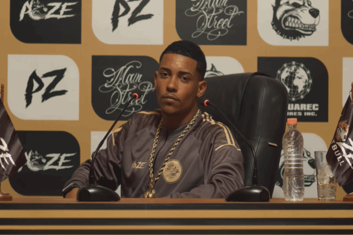 MC Poze do Rodo com expressão séria durante pronunciamento após sofrer roubo milionário. O cantor veste jaqueta cinza e cordão de ouro, sentado atrás de uma bancada com microfones e logomarcas ao fundo.