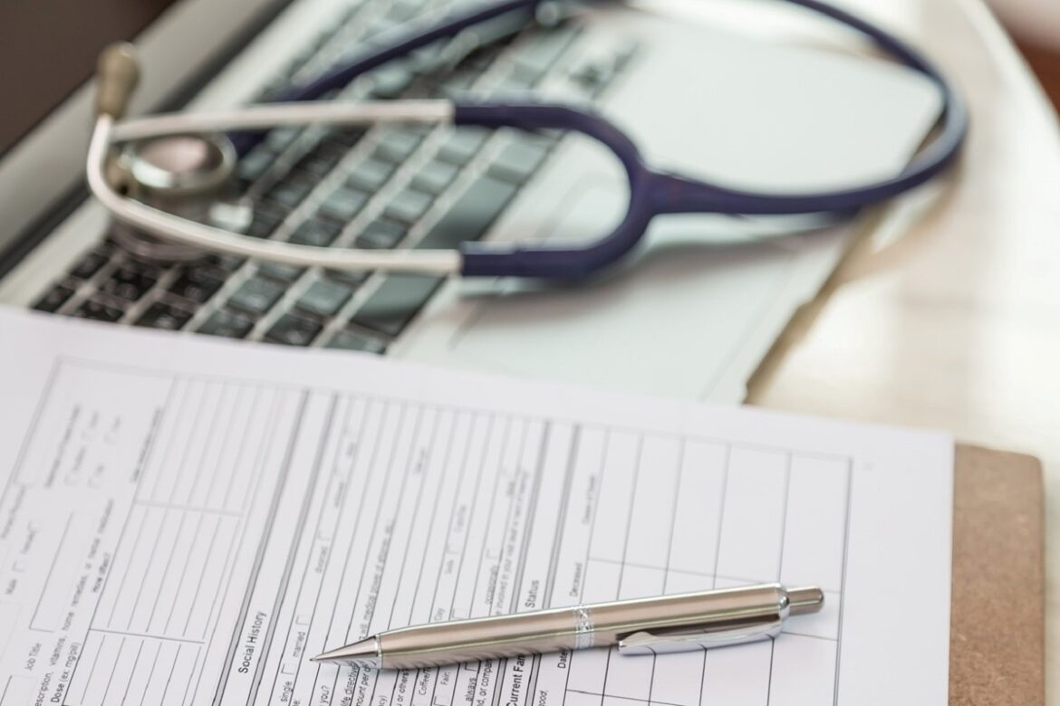 Estetoscópio azul sobre teclado de notebook com caneta prateada e formulário médico em mesa de consultório