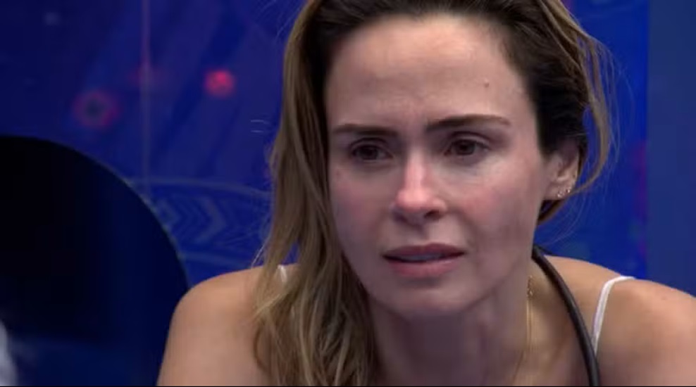 BBB 26: Após saber da morte do pai, Ana Paula Renault decide continuar no programa; entenda o que aconteceu 6 Ana Paula Renault emocionada, com olhos marejados e expressão de dor, cabelo loiro preso, fundo azul da casa do BBB