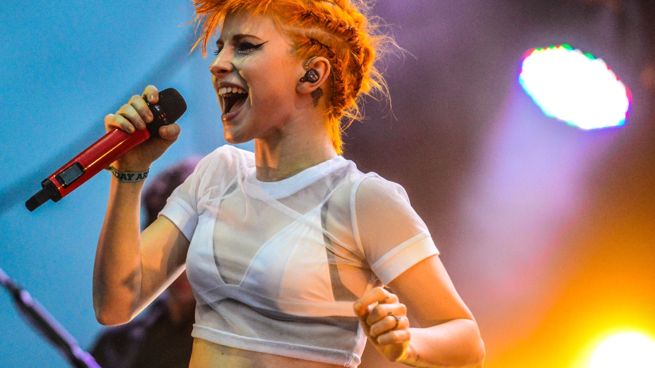 Cantora Hayley Williams vem a São Paulo com show solo; pré-venda começa em breve 6 Hayley Williams, com cabelo laranja curto, canta segurando um microfone vermelho em palco com luz azul e roxa
