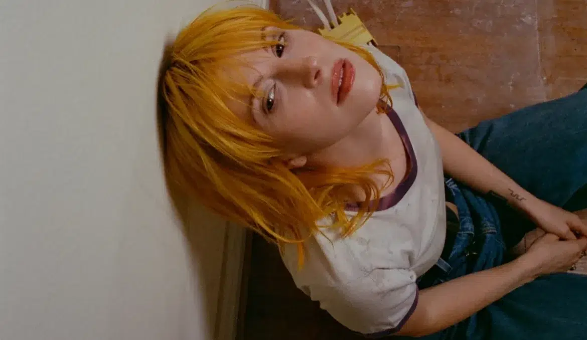 Hayley Williams com cabelo laranja vibrante, camiseta branca e macacão jeans azul, olhando para a câmera.