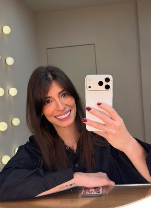 Semelhança com a estrela de "O Diabo Veste Prada" faz brasileira virar sensação na internet 6 Selfie no espelho da influenciadora Emily Estevem Rolim, que viralizou como sósia de Anne Hathaway. Ela sorri, veste uma camisa preta e segura um smartphone branco com três câmeras. Ao fundo, luzes de camarim estão visíveis. Uma tatuagem de corações é vista no pulso e um QR Code na bancada.