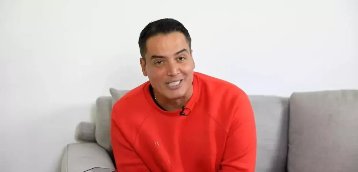 Jornalista Léo Dias usando moletom laranja, sentado em sofá cinza com microfone de lapela