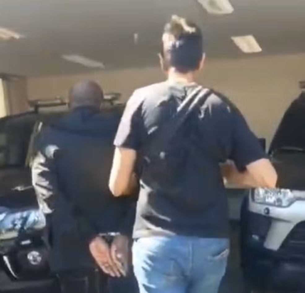 Filho é preso em velório por matar mãe e decepar dedo para movimentar conta bancária 6 Policiais conduzem homem algemado de camiseta azul e calça jeans durante prisão no interior de São Paulo