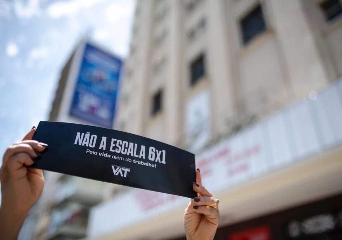 Mãos femininas com unhas pretas erguem cartaz preto do Movimento VAT com a frase "Não à escala 6x1 - Pela vida além do trabalho" em rua