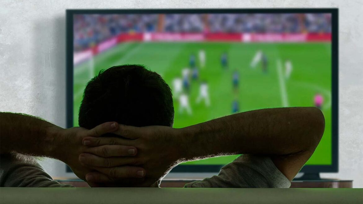 Homem de costas com mãos atrás da cabeça assistindo a jogo de futebol em televisão grande