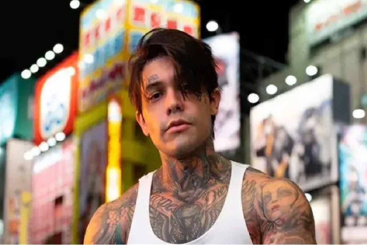 Caso inusitado: influencer é expulso de avião por pegadinha com 'pum' e som 6 Yeferson Cossio com cabelos escuros e tatuagens visíveis veste regata branca à noite diante de letreiros luminosos