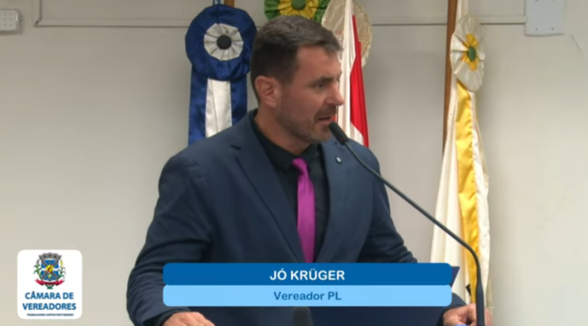 Um homem de meia-idade, com cabelos curtos e barba, usando um terno azul-marinho, camisa preta e uma gravata fúcsia, fala ao microfone em uma tribuna. Ao fundo, há três bandeiras desfocadas e um banner azul e branco no canto inferior esquerdo com o texto "CÂMARA DE VEREADORES". Na parte inferior direita, há um letreiro azul com o nome "JÓ KRÜGER" e "Vereador PL".