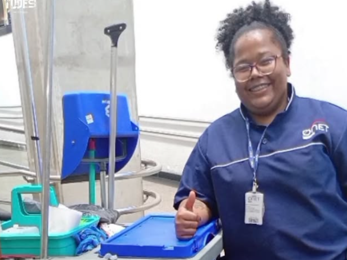 Diarista é imobilizada pela PM na Avenida Paulista após discussão sobre pagamento; filha de 7 anos presenciou a cena 2 Mulher negra sorridente, Jussara Bonfim Silva, usando óculos de grau, aparelho ortodôntico e o uniforme azul de uma empresa de serviços. Ela faz um sinal de