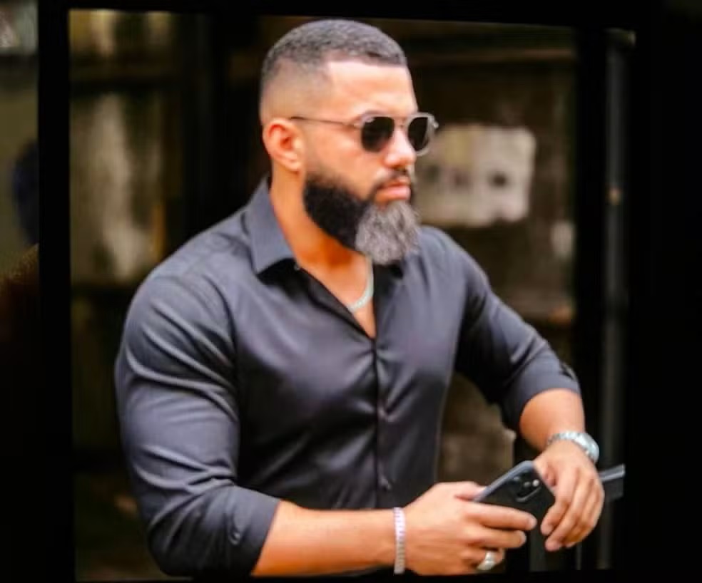 Motorista é morto após jogar carro contra assaltantes para proteger vítima 6 Homem de barba grisalha, óculos escuros e camisa cinza-chumbo segura smartphone, usando relógio e pulseira