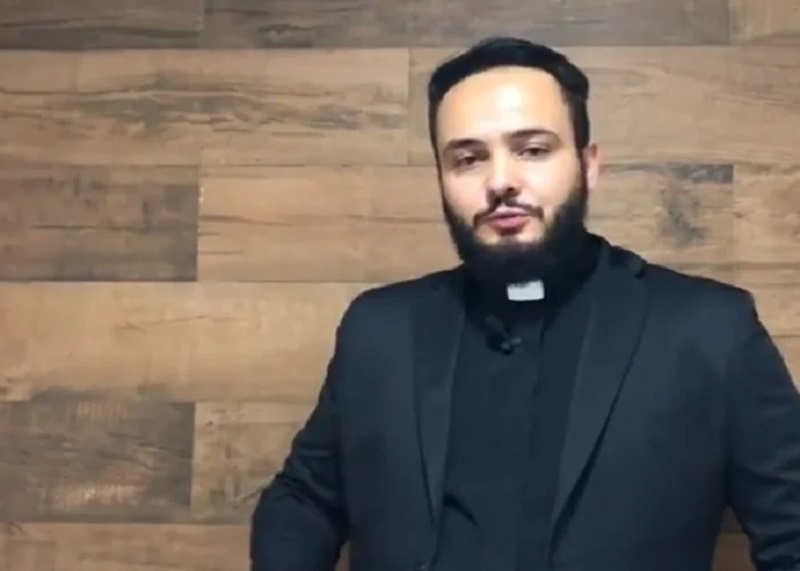 Padre afirma que comer peixe na sexta-feira santa é errado; assista 6 Homem de barba e cabelos escuros, vestindo batina preta, diante de parede de madeira clara, olhando para a câmera